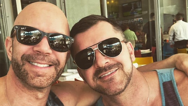 Deux hommes souriants assis sur une terrasse. Ils sont en camisoles et portent des lunettes fumées.