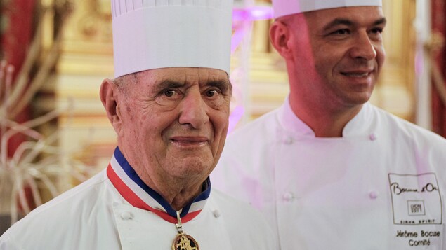 Paul Bocuse est coiffé d'une toque blanche. À son cou,  il porte sa médaille de Meilleur ouvrier de France. 