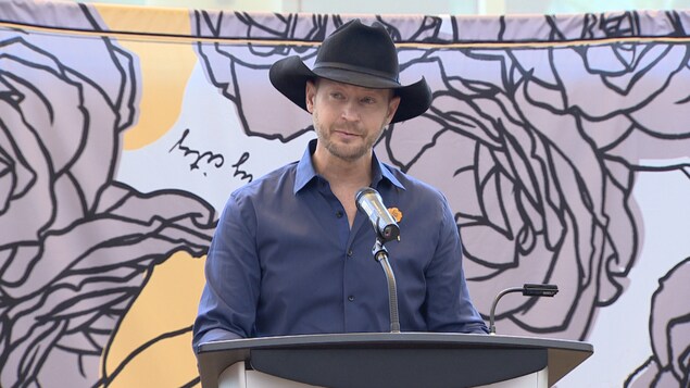 Le chanteur Paul Brandt fait une allocution devant une estrade avec micro. Derrière lui, une grande toile avec le logo de sa campagne de sensibilisation (des roses) est affichée.