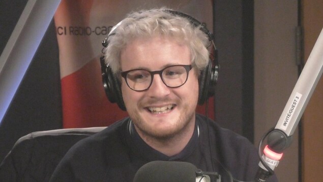 Dans un studio de Radio-Canada, un jeune homme blond avec des lunettes noires sourit en regardant en biais. Il a un casque sur les oreilles et un micro devant la bouche. Dans un studio de Radio-Canada, un jeune homme blond avec des lunettes noires sourit en regardant en biais. Il a un casque sur les oreilles et un micro devant la bouche.