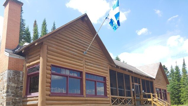 Vue sur le pavillon du lac à L’Épaule. Il s’agit d’un bâtiment en rondins muni d’une cheminée faite de pierres et de briques. Un drapeau du Québec orne la façade. Vue sur le pavillon du lac à L’Épaule. Il s’agit d’un bâtiment en rondins muni d’une cheminée faite de pierres et de briques. Un drapeau du Québec orne la façade.