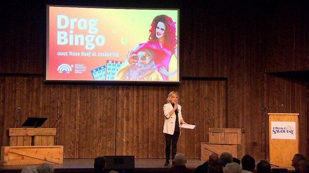 Monique Poirier sur scène devant un public au Pays de la Sagouine. Derrière elle, un immense écran qui porte la mention drag bingo avec Rose Beef et Joséphine.