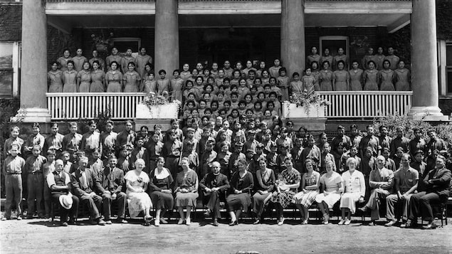 Une foule de jeunes des Premières Nations se tiennent les bras croisées dans le dos pour la photo, leur professeurs blancs assis à l'avant