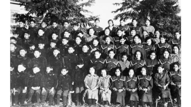 Une vieille photo en noir et blanc de jeunes écoliers autochtones en uniformes, assis en rangées.