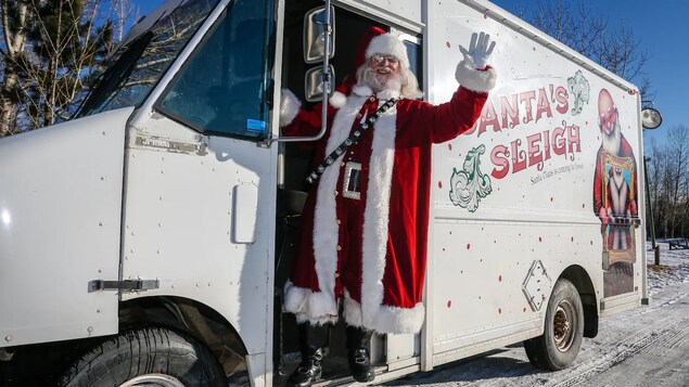 Un père Noël se trouve dans un camion-restaurant. Souriant, il salut la caméra. Sur le camion est peint un père Noël et les mots traîneau du père Noël.