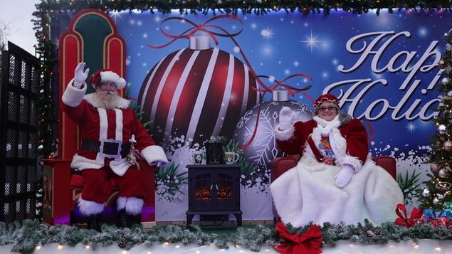 Le père et la mère Noël assis dans leur fauteuil près d'un foyer. Ils envoient la main aux passants.