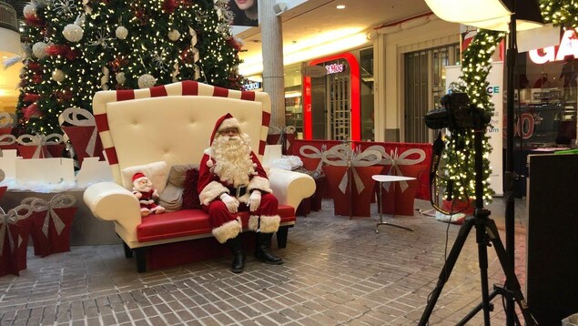 Le père Noël est assis sur un petit sofa de deux places, entouré d'un décor de Noël. Devant lui est installé une appareil photo.