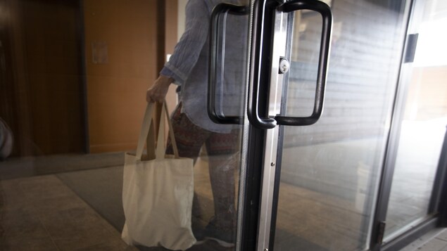 Une personne de dos, portant un sac à la main, derrière une porte de verre.