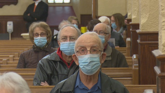 Des gens sont photographiés de face alors qu'ils sont assis à l'église avec des masques bleus au visage.