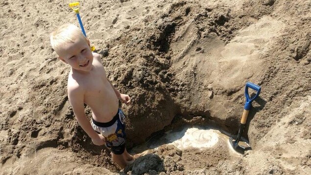 Un petit garçon en maillot de bain se tient debout dans un trou creusé dans le sable. Il regarde l'objectif. Deux pelles sont plantées dans le sable. Un petit garçon en maillot de bain se tient debout dans un trou creusé dans le sable. Il regarde l'objectif. Deux pelles sont plantées dans le sable.