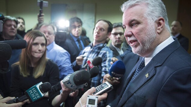 Le premier ministre Philippe Couillard est entouré de journalistes qui pointent vers lui leurs micros et leurs caméras.