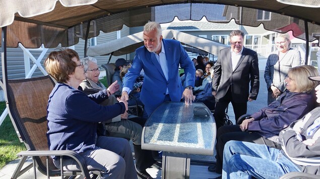 Philippe Couillard serre la main d'une femme qui est assise, avec d'autres personnes, dans une chaise extérieure berçante.