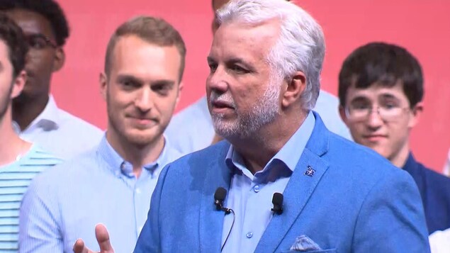 Philippe Couillard s'exprime sur une scène, des jeunes se tiennent derrière lui.