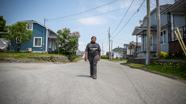 Une jeune femme vêtue d'un t-shirt gris avance au milieu d'une rue résidentielle bordée de maisons.