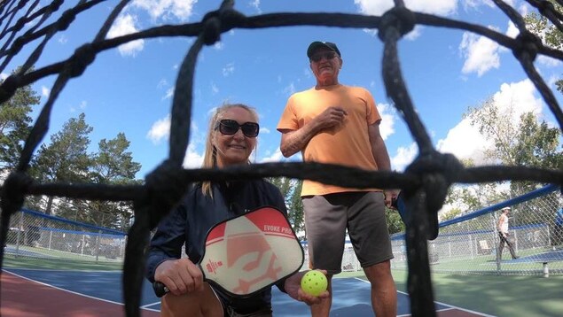 Plan vue d'en dessous d'une femme agenouillée et un homme se tenant debout devant un filet de Pickeball au parc Father Basil Markle de Saskatoon.