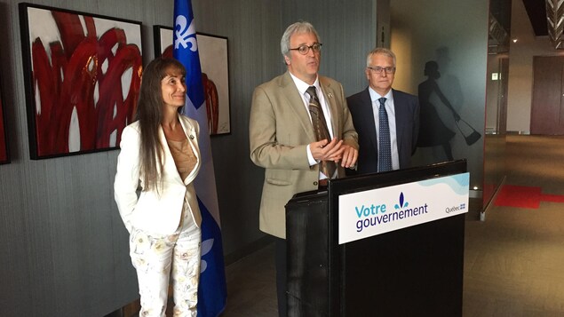 Pierre Dufour, encadré d'une femme et d'un homme, parle derrière un lutrin du gouvernement du Québec.