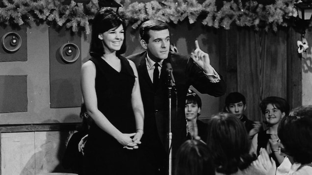 Dans une salle, la chanteuse Michèle Richard se tient à côté de l'animateur Pierre Lalonde qui parle au public. Celui-ci applaudit.
