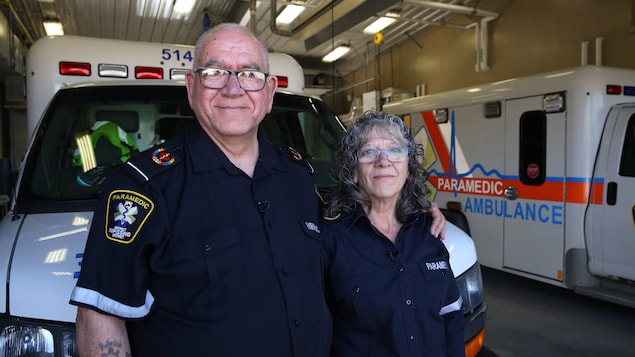 Pierre et Marilyne sourient à la caméra dans un garage avec des ambulances derrière eux.