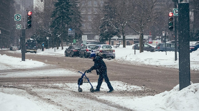 Un piéton traverse la rue en s'appuyant sur son déambulateur, dans une rue enneigée du centre-ville de Montréal.