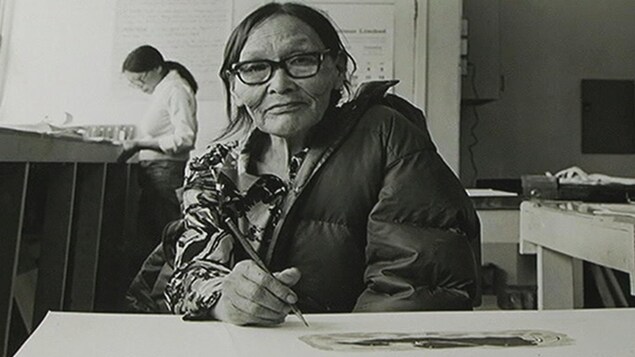 Une femme inuite, vêtue d'un manteau, semble prendre une pause de son dessin le temps que le photographe prenne la photo. Elle porte des lunettes à forte monture et tient un crayon en main. Une femme inuite, vêtue d'un manteau, semble prendre une pause de son dessin le temps que le photographe prenne la photo. Elle porte des lunettes à forte monture et tient un crayon en main.
