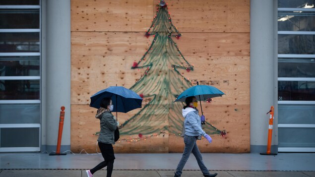 Deux femmes avec une parapluie marchent sur un trottoir avec un dessin de sapin de Noël au mur.
