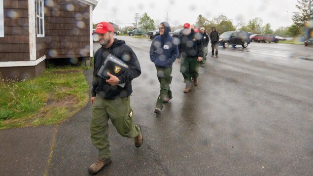 Des pompiers américains marchent à la queue leu leu, souriant, sous la pluie, dans un stationnement. 