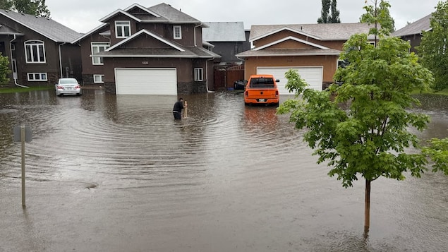 Une rue résidentielle inondée sous la pluie à Brandon. Un homme se tient avec de l'eau jusqu'aux genoux au milieu de la chaussée inondée, devant des maisons de banlieue et une camionnette orange.
