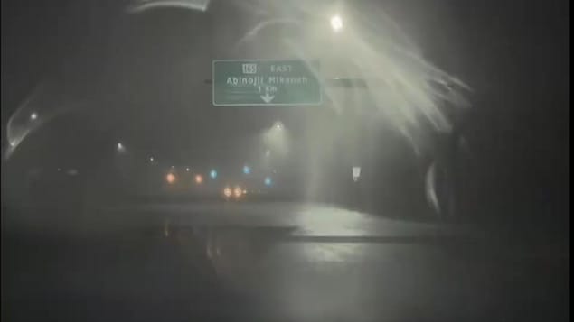 Une vue de nuit à travers un pare-brise flou par la pluie montre un panneau routier vert indiquant la route 165 Est Abinoojii Mikanah à 1 kilomètre, sous des lampadaires enveloppés de brume.