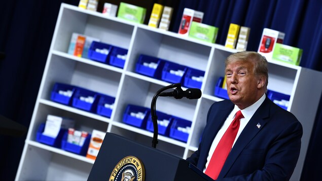 M. Trump est au micro à côté d'étalages de médicaments. Ces derniers ont l'air en pente afin d'illustrer la baisse des prix voulue par le président américain.