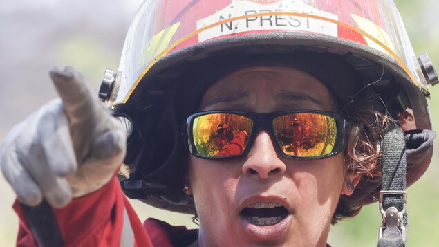 Une pompière casquée, en uniforme, pointe devant elle. Dans ses lunettes fumées, on voit d'autres pompiers devant un feu de forêt.