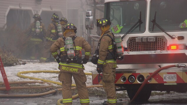 Des pompiers à côté d'un camion devant une résidence de laquelle sort de la fumée Des pompiers à côté d'un camion devant une résidence de laquelle sort de la fumée