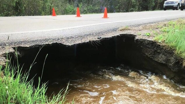 De l'eau s'écoule sous une route dont le bord est effondré. La route est bloquée par trois cônes oranges. De l'eau s'écoule sous une route dont le bord est effondré. La route est bloquée par trois cônes oranges.