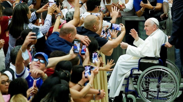 Pope Francis in his wheelchair greets a crowd of people taking pictures with their phone.