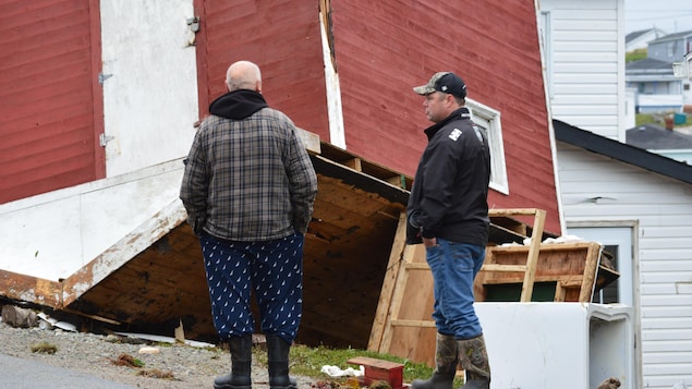 Deux hommes devant une grosse cabane à l'envers après avoir été renversée par un ouragan.