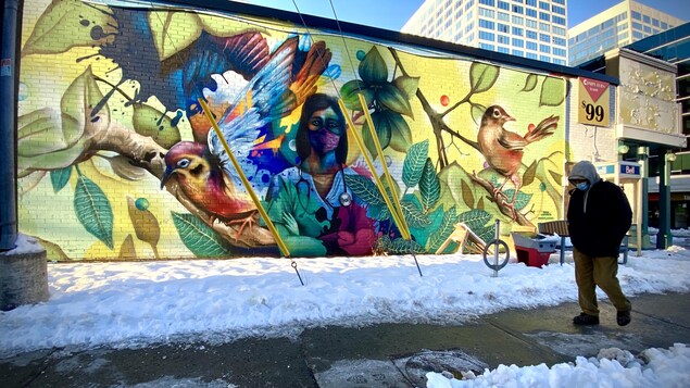 Un homme masqué passe devant une murale colorée illustrant une infirmière portant le masque accompagnée d'oiseaux et de feuilles d'arbres.