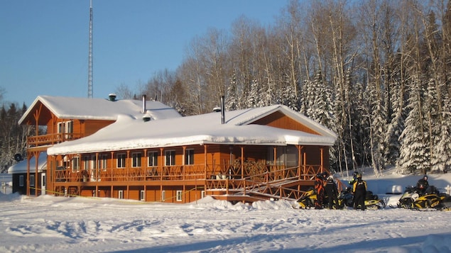 Un chalet de bois en hiver. Des motoneigistes discutent autour de leur motoneige.