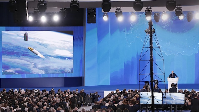 Un écran géant montre les images d'une simulation du trajet du nouveau missile nucléaire russe alors que Vladimir Poutine s'adresse à l'assemblée fédérale debout derrière un lutrin.