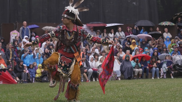 Un homme, vêtu d'une tenue de danse traditionnelle autochtone, incluant une coiffe à plumes et des vêtements décorés de motifs complexes et de franges, est en mouvement sur un terrain herbeux. Il tient un tissu rouge dans sa main droite. En arrière-plan, une foule de personnes, dont beaucoup tiennent des parapluies, assiste à l'événement, suggérant un pow-wow sous la pluie.