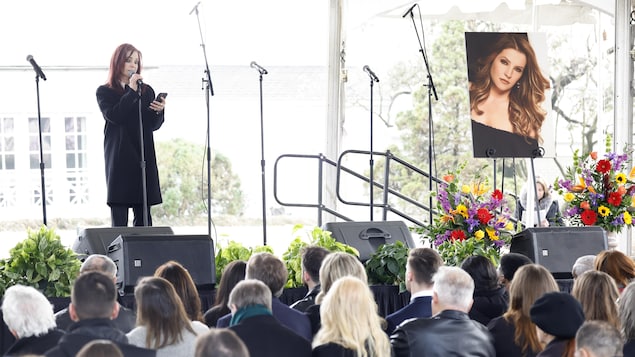 Une femme sur scène parle dans un micro, devant une foule assise lors d'un office funèbre. 