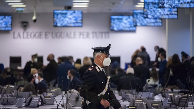 Un policier italien traverse la salle d'audience. Des gens discutent en arrière-plan.