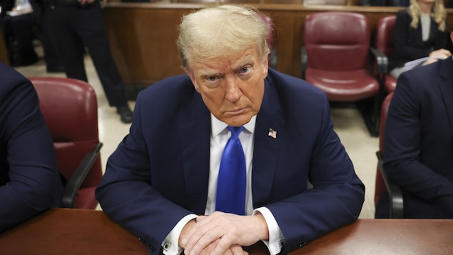 Assis au banc des accusés, Donald Trump, mains jointes, regarde devant lui.