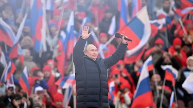 Nakataas ang mga kamay ni Vladimir Putin habang binabati ang audience, iwinawagayway ang mga bandila ng Russia sa kanyang likuran.
