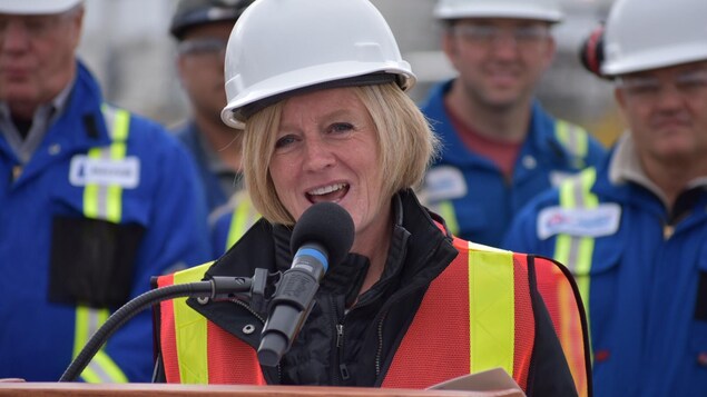 La première ministre Albertaine Rachel Notley parle dans un micro avec des travailleurs en blouse bleue derrière elle. La première ministre Albertaine Rachel Notley parle dans un micro avec des travailleurs en blouse bleue derrière elle.