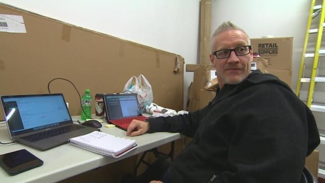 Assis à une table sur laquelle se trouvent deux ordinateurs portables et parmi des cartons vides, un homme regarde derrière lui, en biais, l'air amusé. Assis à une table sur laquelle se trouvent deux ordinateurs portables et parmi des cartons vides, un homme regarde derrière lui, en biais, l'air amusé.
