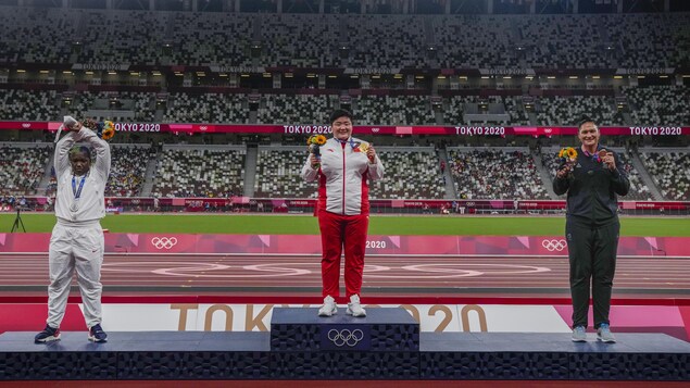 Raven Saunders, sur la seconde marche du podium, croise les bras au-dessus de sa tête alors que deux autres femmes posent pour la caméra en montrant leur médaille dans un stade.