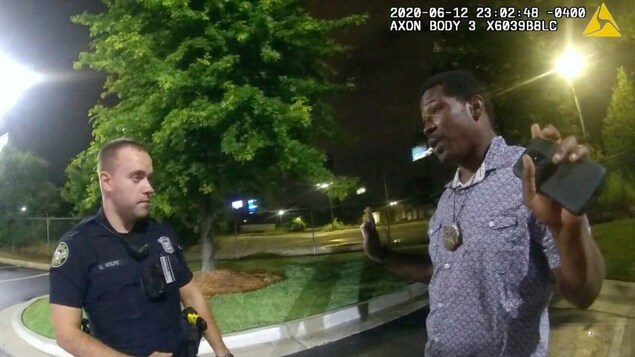 Un homme noir parle les bras levés, un téléphone portable dans une main, face à un policier.