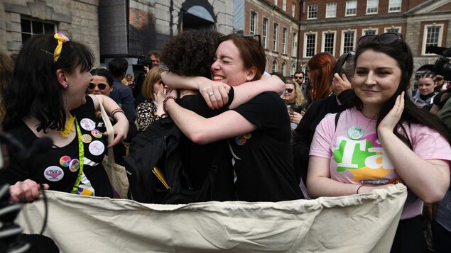 Deux femmes, l'une frisée, l'autre non, s'enlacent. Celle de gauche sourit, entourée par la foule.