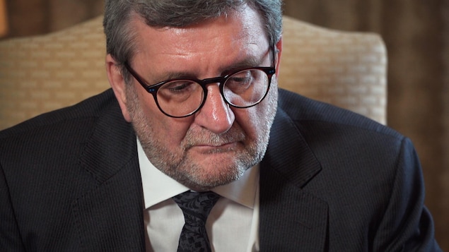 Régis Labeaume avec des lunettes, un veston et une cravate, le regard sérieux, regarde vers le bas