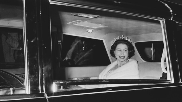 Une jeune reine Élisabeth II sourit en regardant par la vitre arrière d'une grande voiture noire.