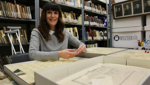 Une femme sourit à la caméra avec des archives derrière elle. Elle tient dans ses mains une petite relique. Une femme sourit à la caméra avec des archives derrière elle. Elle tient dans ses mains une petite relique.
